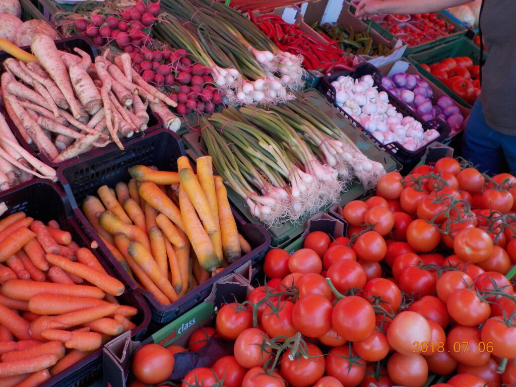This picture shows a vegetable stand at an open-air market: featured are tomatoes, carrots, green onions, radishes, horseradish, garlic, and peppers.