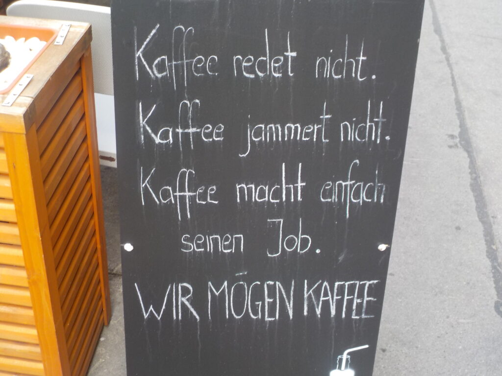 This picture shows chalkboard in front of a café that reads "Kaffee redet nicht. Kaffee jammert nicht. Kaffee macht einfach seinen Job. Wir mögen Kaffee."