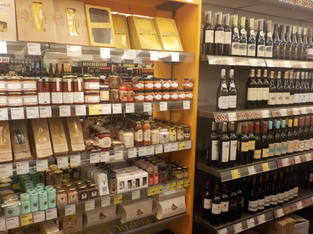 This picture shows the shelves of a gourmet food store, which feature olives, sauces, other condiments, and a variety of wines.