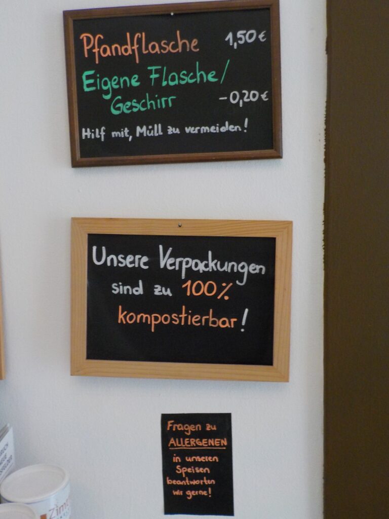 This picture shows three signs in a vegan cafe that communicate their efforts to be sustainable: there is a deposit on all bottles and a discount for any customer who brings their own packaging; the café's packaging is compostable, and the employees gladly answer questions about allergens.