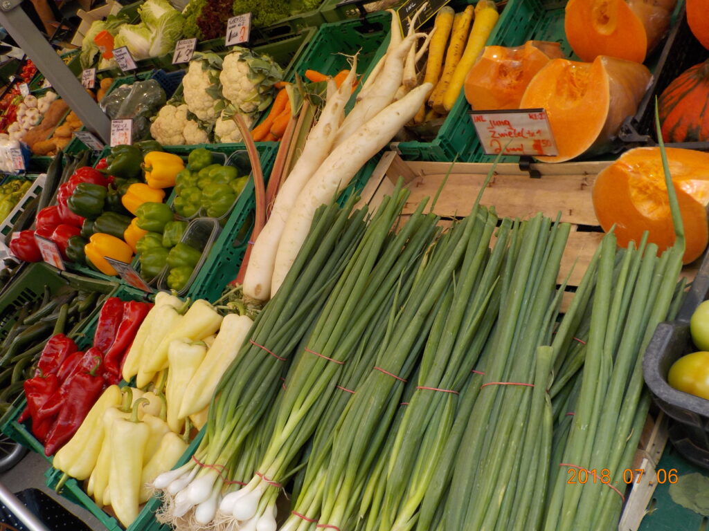 This picture shows a market stand with green onions, radishes, assorted peppers, cauliflower, and other vegetables.