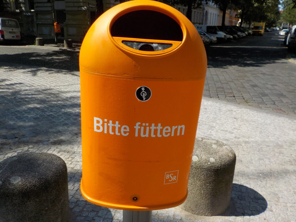 This picture shows an organ trash can in the city bearing the words "bitte füttern" on it.