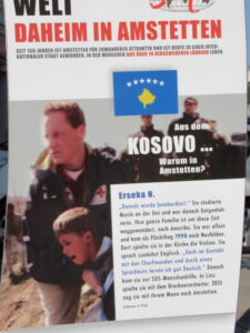 "Menschen aus aller Welt daheim in Amstetten" poster: full text of image is included as the reading texts for Kosovo below. An image of the Kosovo flag and a Red Cross worker helping a young crying boy.